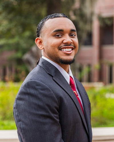 A professional headshot of a smiling young man with a goatee and braided hair, wearing a dark grey suit and a red tie, with a blurred campus building and greenery in the background.