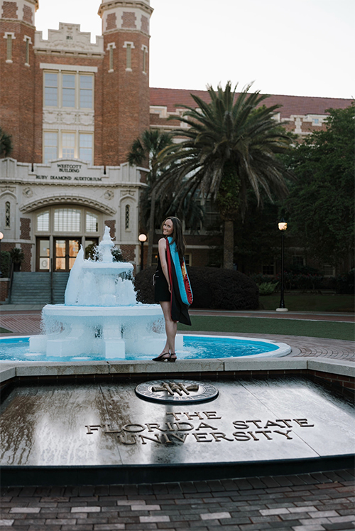 A graduate wearing a black academic gown with a bright blue and red hood stands smiling behind a turquoise fountain, with the Florida State University Westcott Building and palm trees in the background.