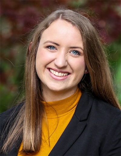 A close-up professional headshot of a smiling woman with long brown hair, wearing a yellow turtleneck and a black blazer against a soft-focus background of green and red foliage.
