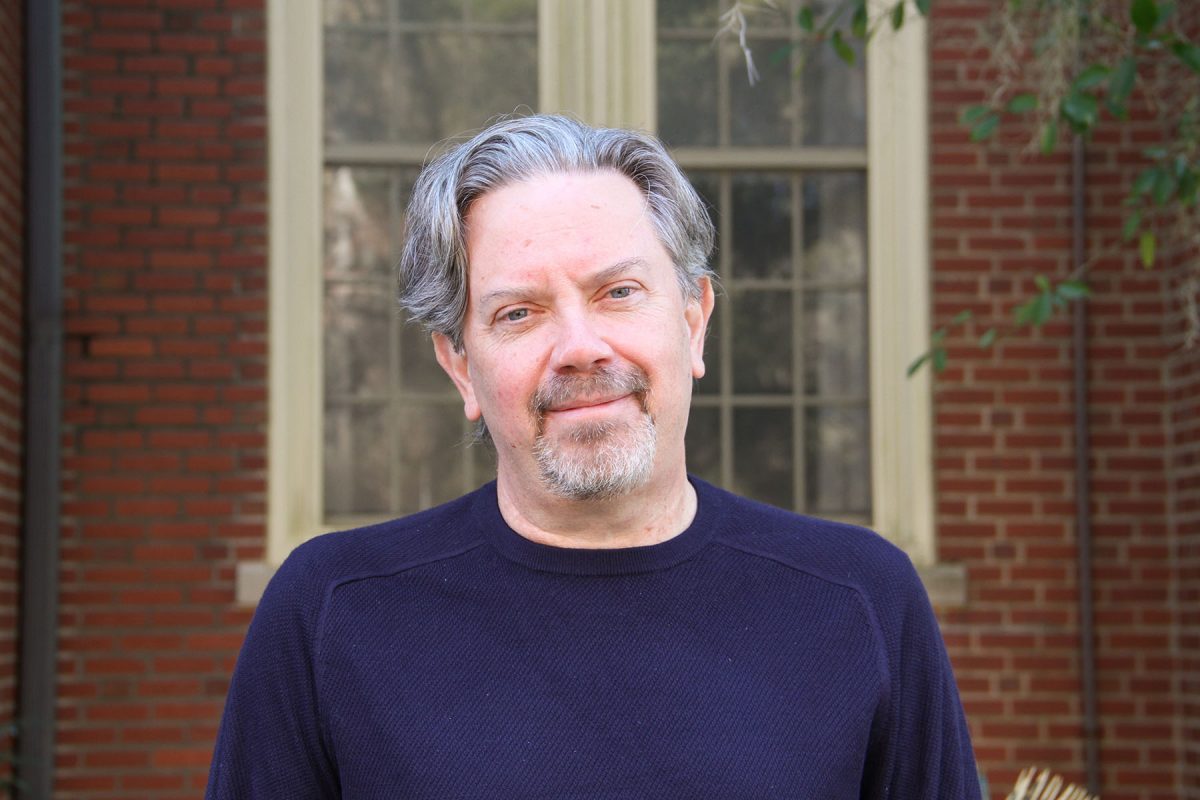 A medium shot of Michael Bishop, a middle-aged man with salt-and-pepper hair and a goatee, wearing a dark navy sweater and smiling slightly in front of a red brick building.