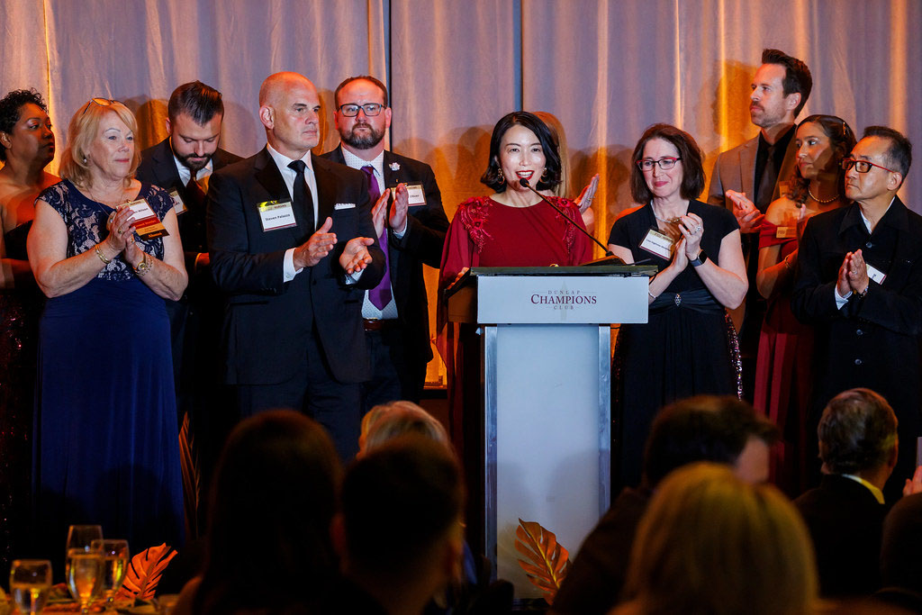 A wide shot of a formal awards ceremony or gala. A woman with dark hair, wearing a red long-sleeved dress, stands at a light blue podium labeled "CHAMPIONS" and speaks into a microphone. To her left and right, several colleagues in suits and formal dresses stand on stage, clapping and looking toward her with supportive expressions. The background features warm, draped gold curtains, and the blurred silhouettes of an audience are visible in the foreground.