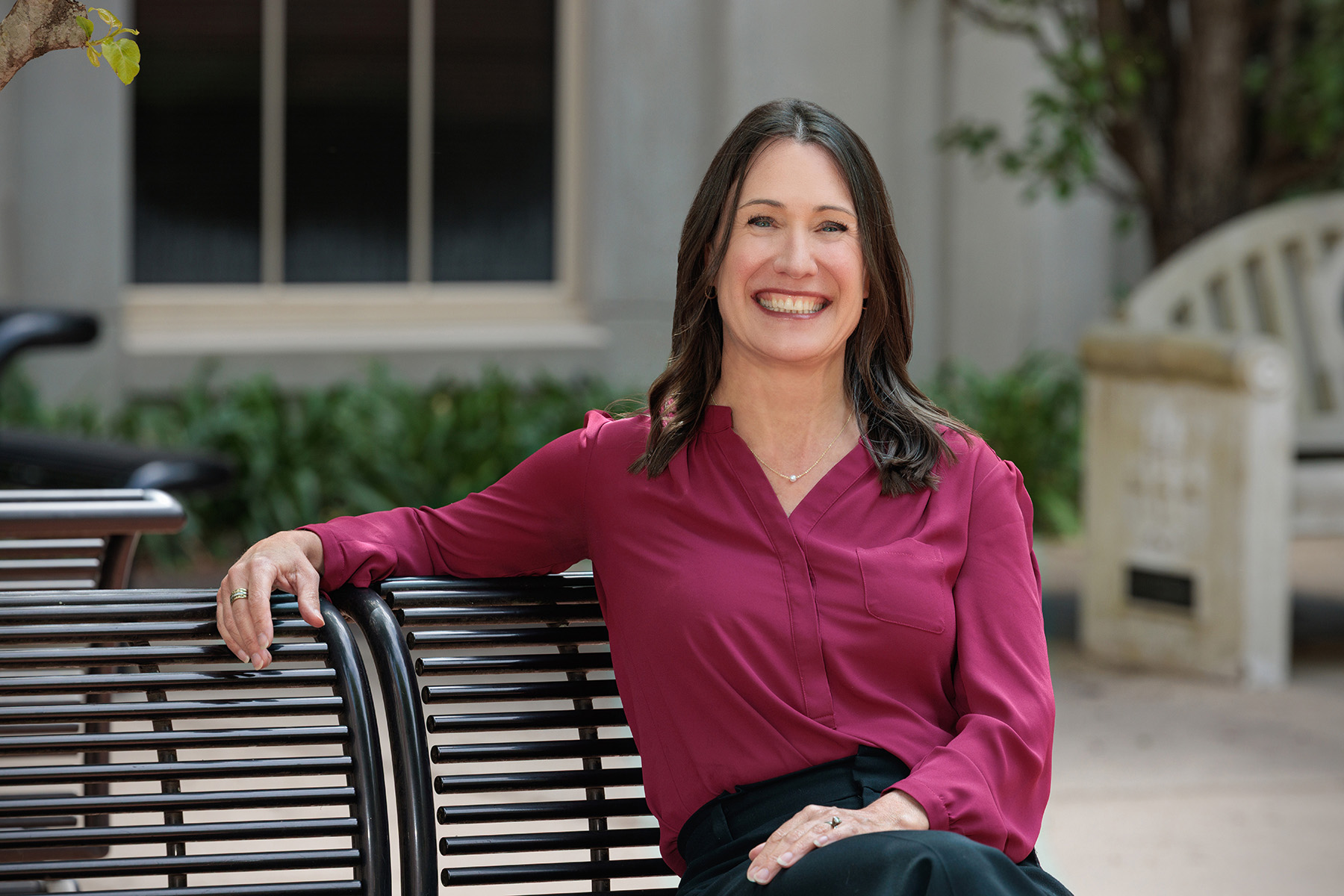 A woman smiles for a photo, sitting at a bench, wearing a burgundy shirt