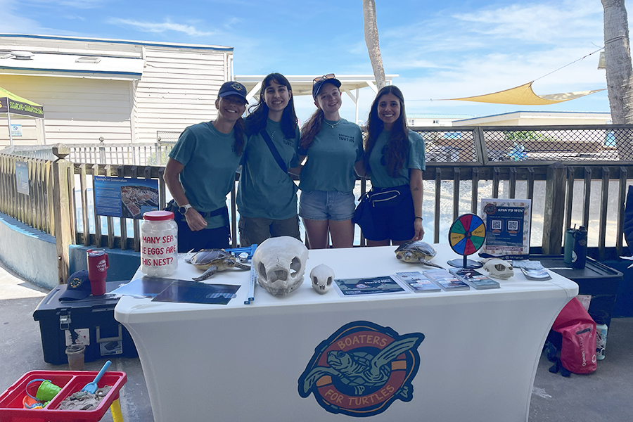 Four people stand at a table with a logo reading “Boaters for Turtles” that includes an image of a sea turtle. Various objects, including sea turtle skulls, a model sea turtle and educational pamphlets, are on the table.
