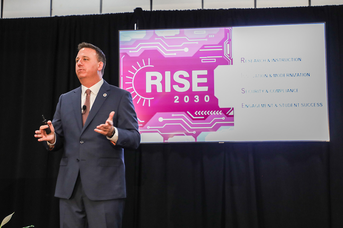Man in a suit talking and standing in front of a video screen that says RISE.