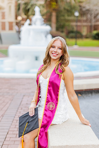 A blonde graduate in a white floral dress and bright pink stole sits on a stone ledge, holding her graduation cap with a turquoise fountain in the background.