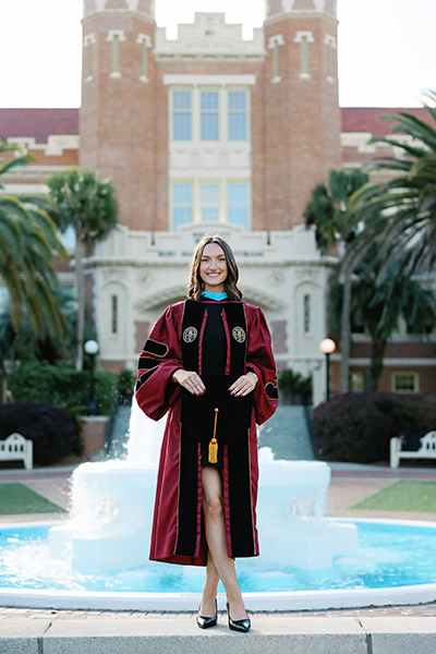 A smiling graduate in a doctoral gown stands in front of a turquoise campus fountain, with a historic brick university building and palm trees in the background.