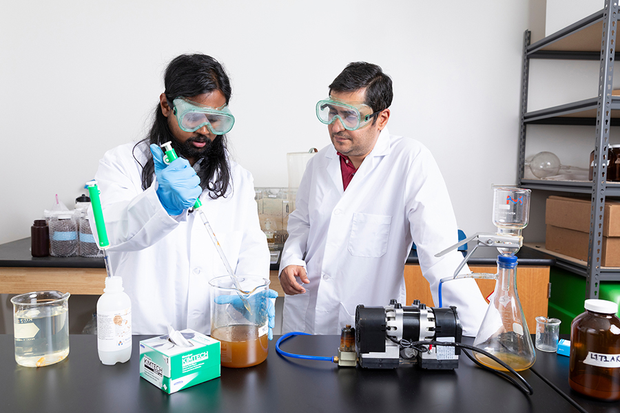 Researcher in safety goggles uses a pipette to transfer liquid into a beaker containing a cloudy brown sample while a colleague observes beside lab equipment.