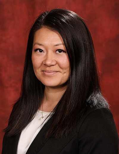A professional headshot of a woman with long dark hair, wearing a black blazer and white blouse with a pearl necklace, smiling against a textured deep red background.