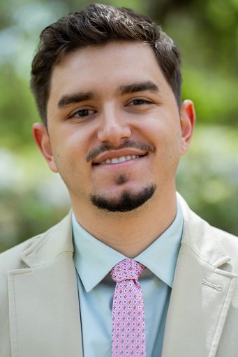 A young man with dark hair and a goatee wearing a tan blazer, a light blue dress shirt, and a patterned pink tie.