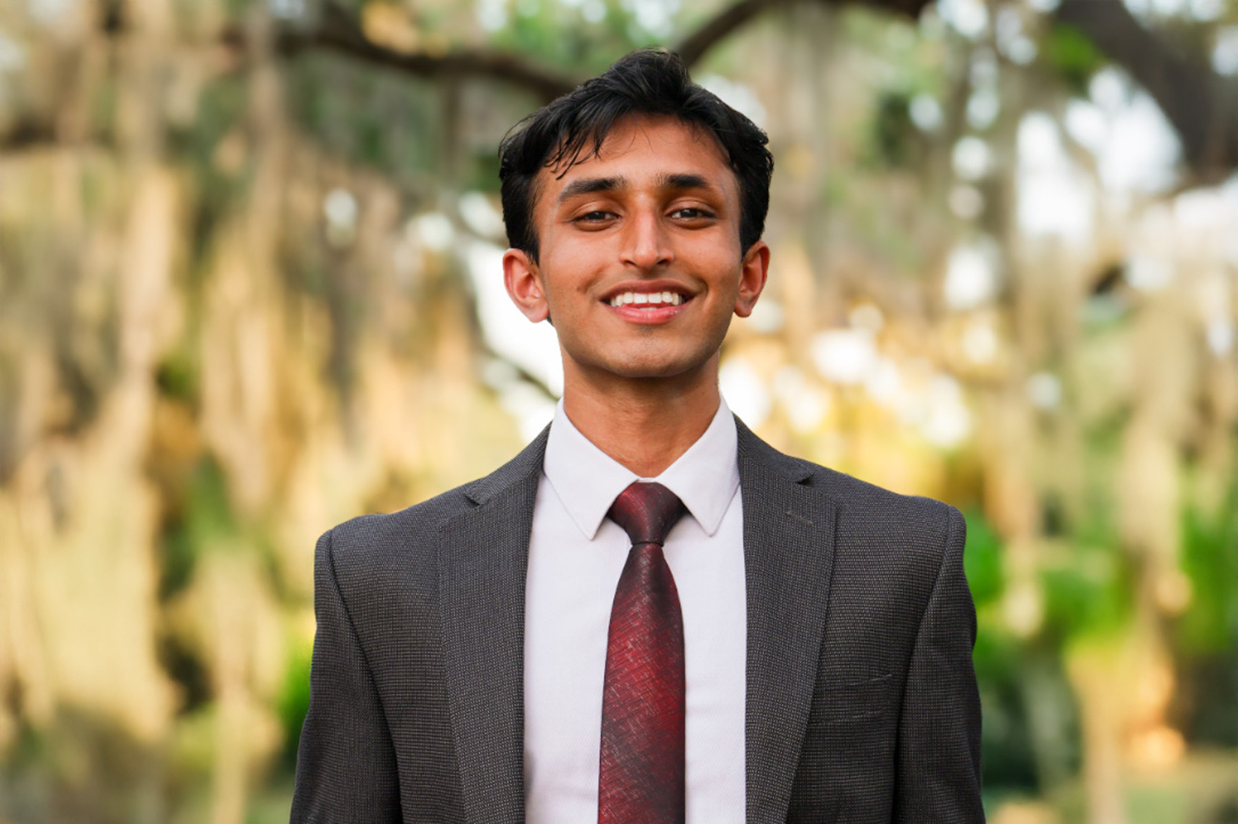 A smiling young man with dark hair and a suit stands outdoors against a blurred background of trees.