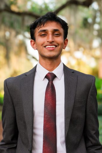 A young man with dark hair and a tan complexion wearing a dark charcoal suit, a white dress shirt, and a solid burgundy tie.