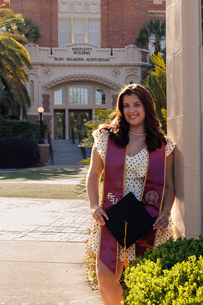 A smiling graduate with brunette hair wears a polka-dot ruffle dress and a FSU stole while holding her cap in front of the Westcott Building on the Florida State University campus.