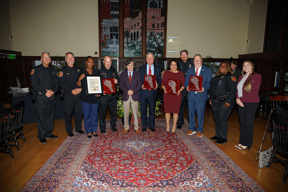 A group of 11 people, including police officers, stand and hold awards in room with wood floors and decorative rug.