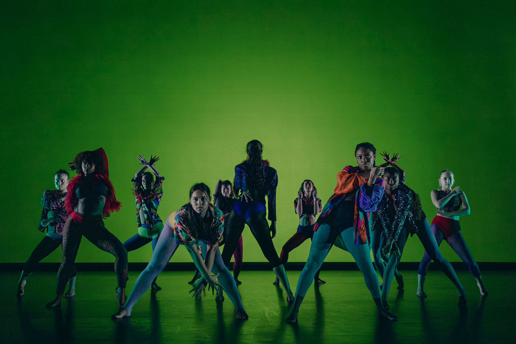 A group of diverse dancers in colorful, eclectic costumes perform an expressive routine against a solid green background.