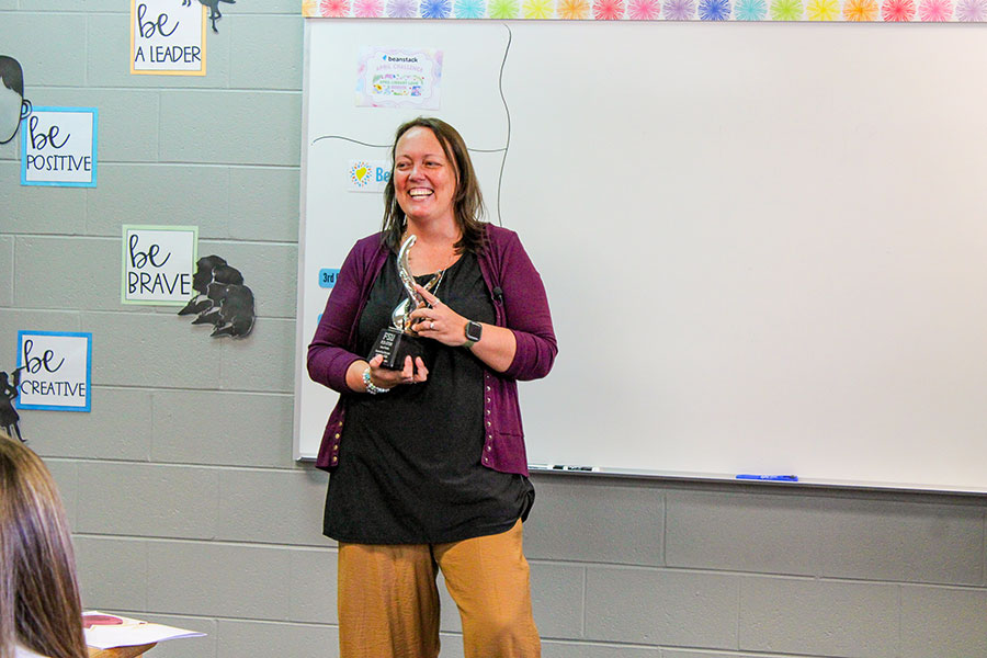 A woman wearing tan pants, a black shirt and purple blouse holding a trophy in a classroom in front of a white board.