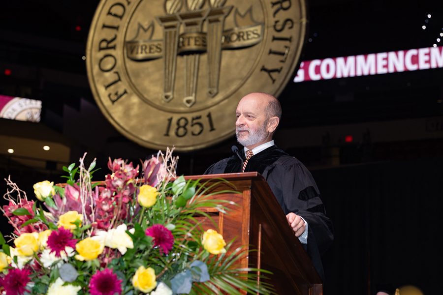 Mark Zeigler addresses graduates from a podium with flowers in front of it in front of the gold FSU seal.