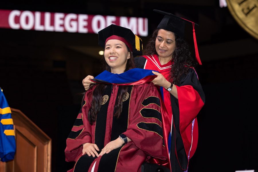 A female asian doctoral graduate smiles while her female faculty mentor places the doctoral hood on her shoulders.
