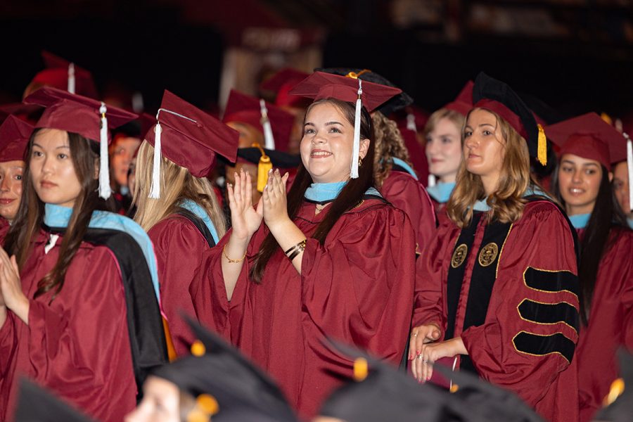 Rows of graduates sit on the ceremony floor wearing maroon caps and gowns.