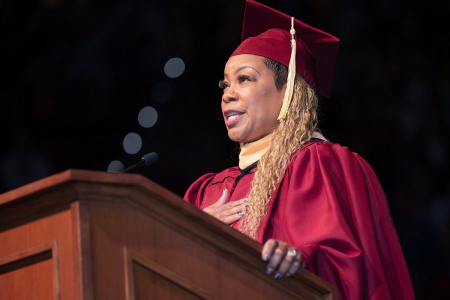 A woman in a garnet graduation cap and gown speaks from a wooden lectern with one hand over her heart during a commencement ceremony.