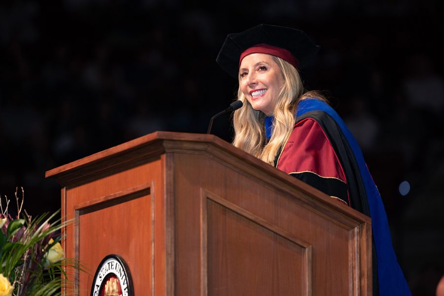 Sarah Blakely smiles while speaking at a wooden lectern during commencement ceremony.