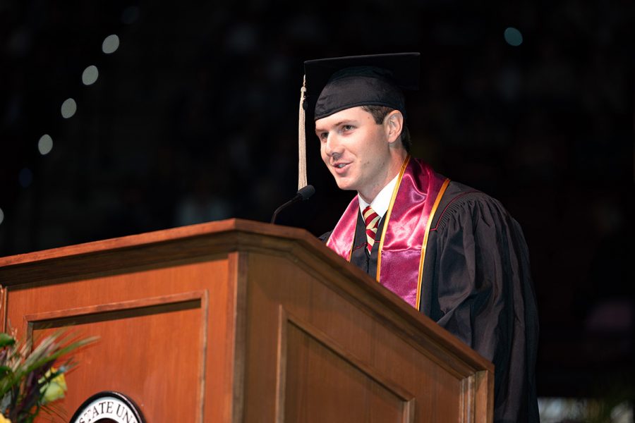 A male graduate wearing a black cap and gown with a garnet stole delivers a speech from a wooden Florida State University podium.