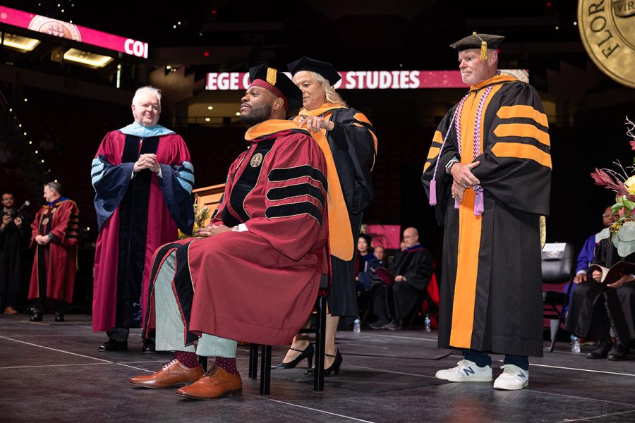A male black doctoral student receives his doctoral hood while sitting on a bench.