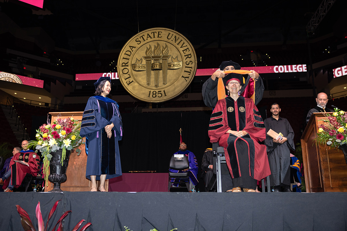The gold FSU seal sits center stage as a female doctoral graduate smiles while sitting on a bench receiving her hood.