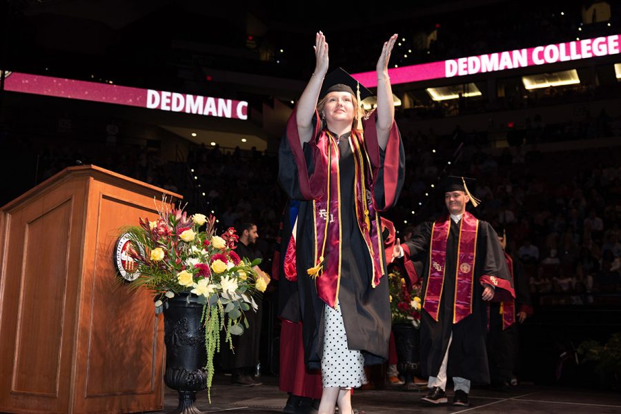 A female graduate in a black gown and garnet stole raises her hands in celebration while walking across the stage during a Florida State University commencement ceremony.