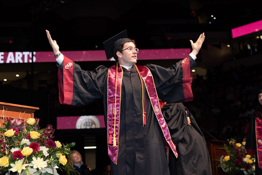 A graduate in a black gown and garnet stole raises his arms in celebration while crossing the stage during a Florida State University commencement ceremony.