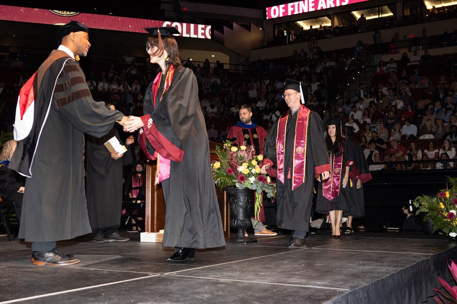 A university official shakes hands with a graduate in a black and garnet gown on stage while other students wait in line during a commencement ceremony.
