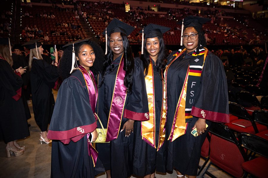 Four Florida State University graduates smile together while wearing their black and garnet academic regalia inside an arena.