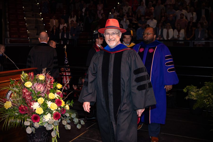 A smiling academic wearing a black doctoral gown and a bright garnet fedora walks across the stage during a Florida State University commencement ceremony.