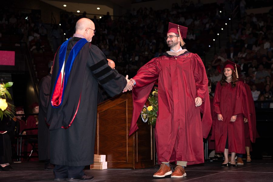 A doctoral graduate in a garnet gown shakes hands with a university official on stage during a graduation ceremony.