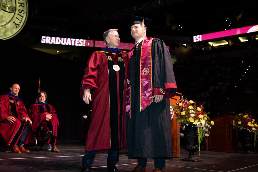 A Florida State University official in academic regalia stands with his arm around a graduate on stage during a commencement ceremony.