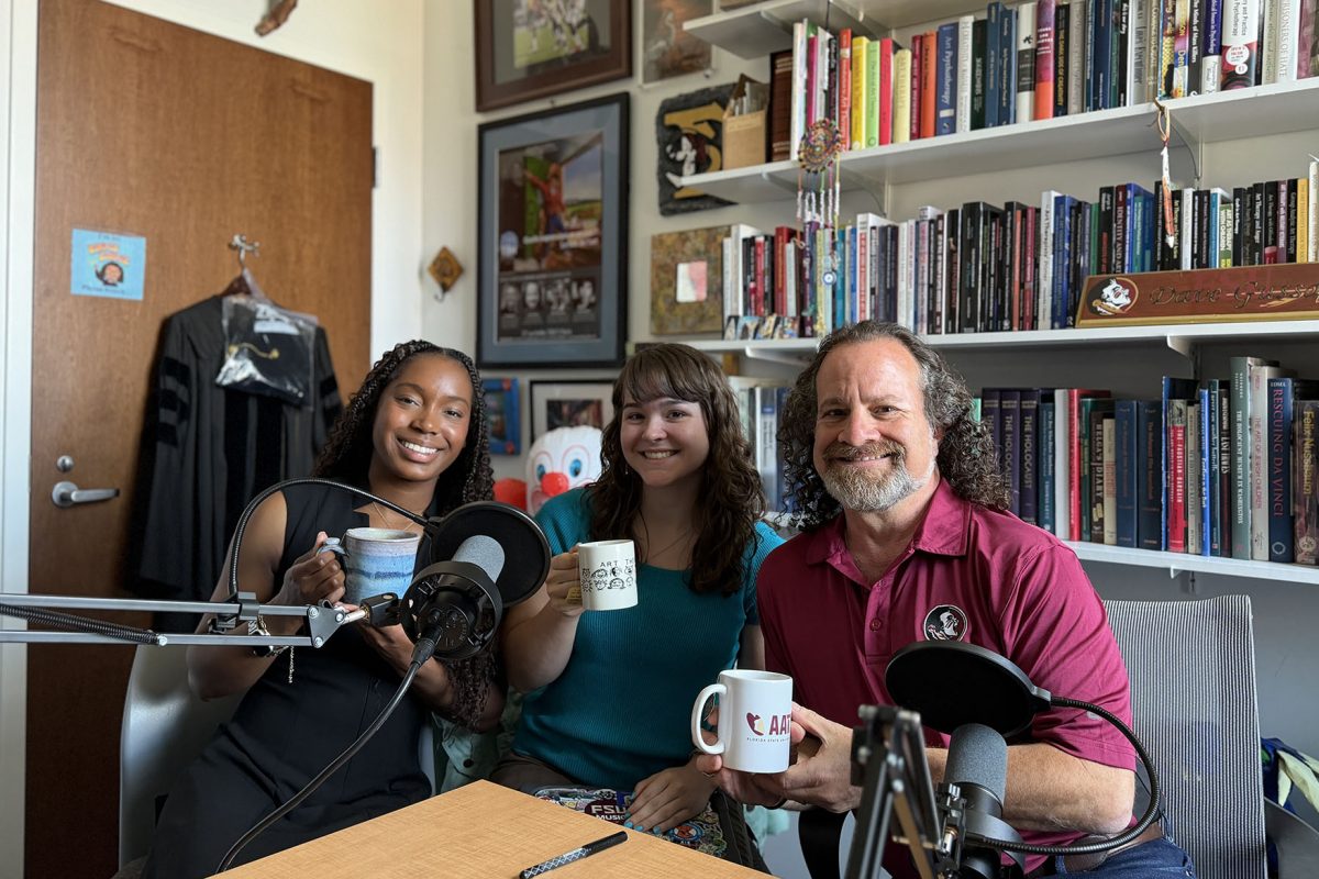 Malea Burroughs, Sydney Nichols and Dave Gussak sit together in a book-lined office recording a podcast at Florida State University. Burroughs and Nichols hold coffee mugs beside podcast microphones while Gussak smiles at the camera.