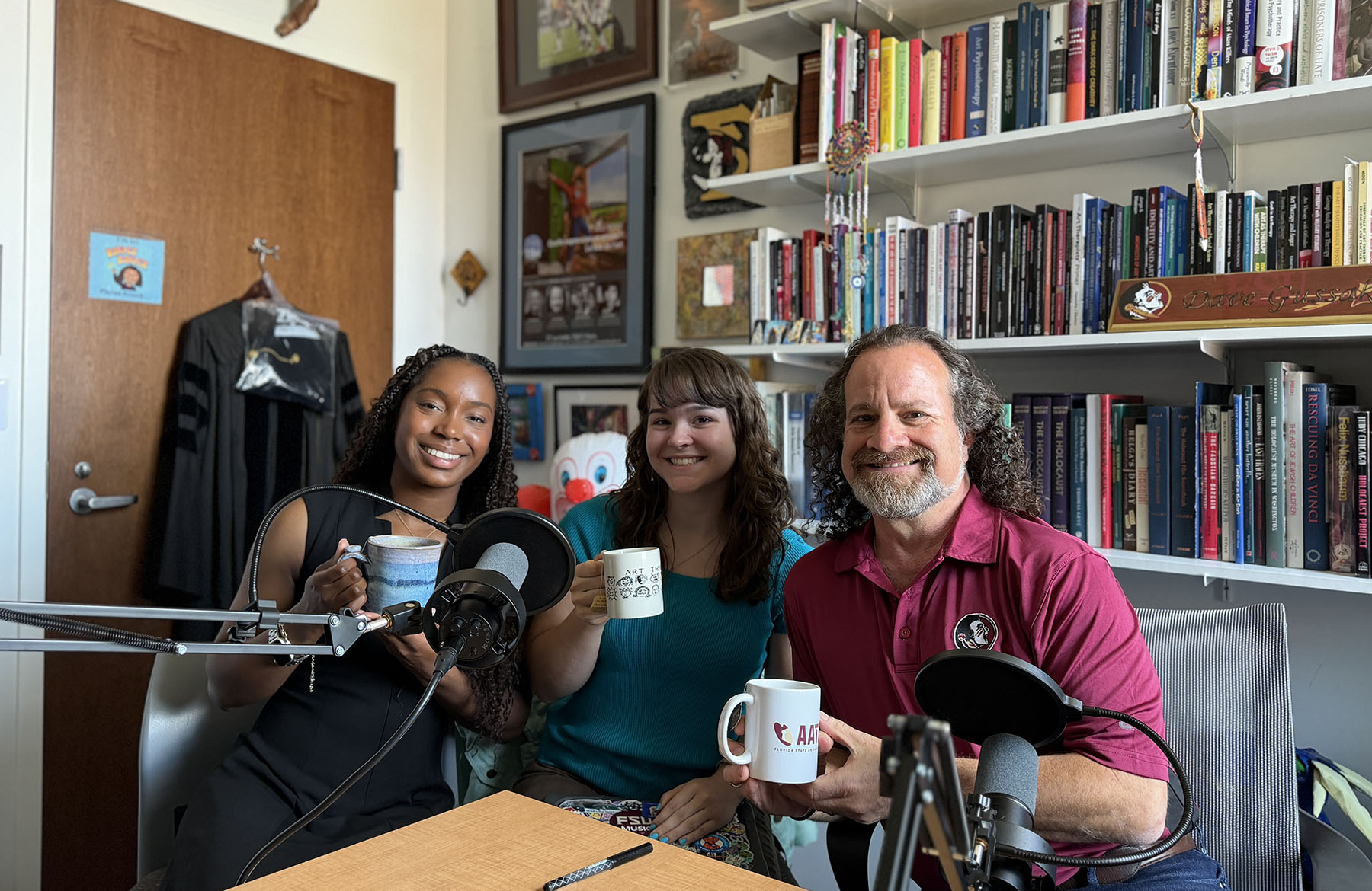 Malea Burroughs, Sydney Nichols and Dave Gussak sit together in a book-lined office recording a podcast at Florida State University. Burroughs and Nichols hold coffee mugs beside podcast microphones while Gussak smiles at the camera.