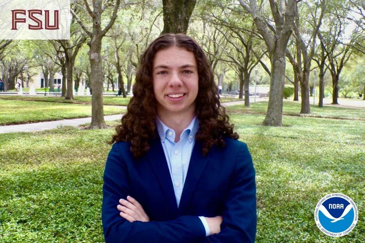 A young man in a light blue shirt and dark blue blazer folds his arms and smiles in front of an outdoor green space