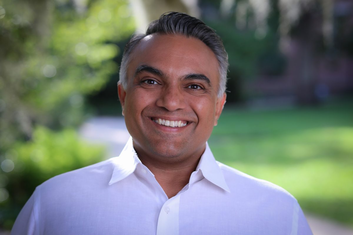 Head-and-shoulders portrait of a person wearing a white button-down shirt, standing outdoors with greenery and campus buildings softly blurred in the background.