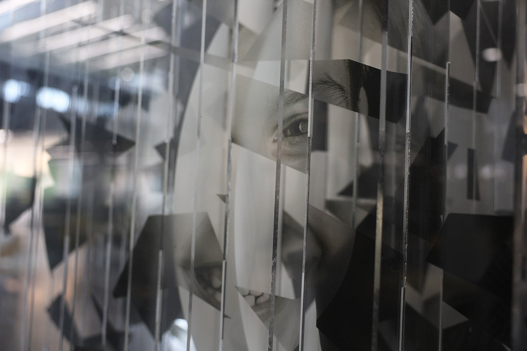 A black-and-white, abstract photograph of a human face fragmented across multiple vertical, transparent panels, revealing a single sharply focused eye amidst sharp, geometric shards.