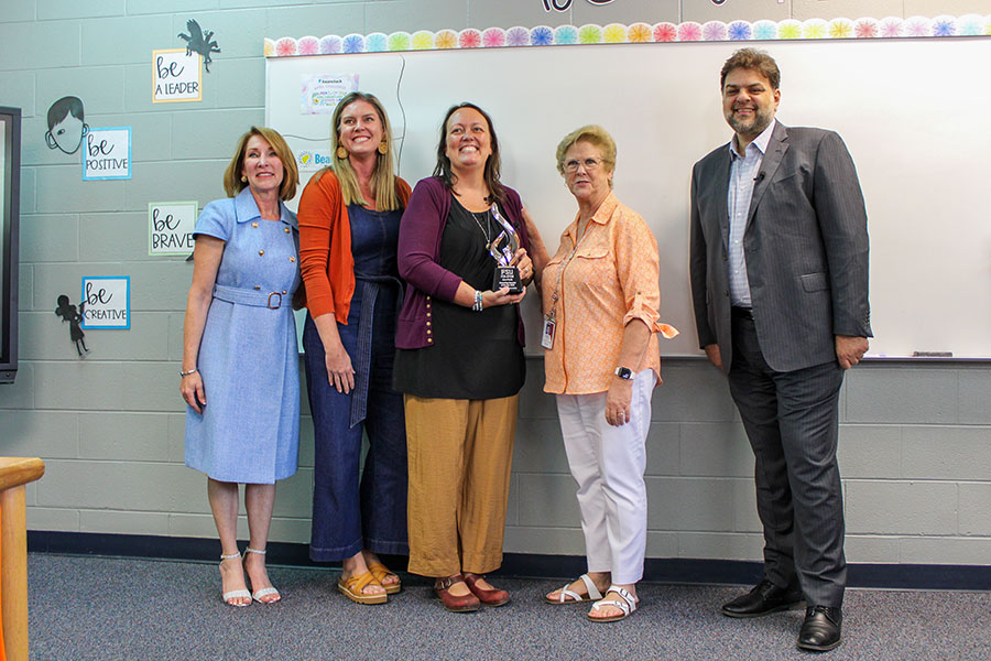 A group of five people post for a photo in an elementary classroom with a woman in the middle holding a trophy.