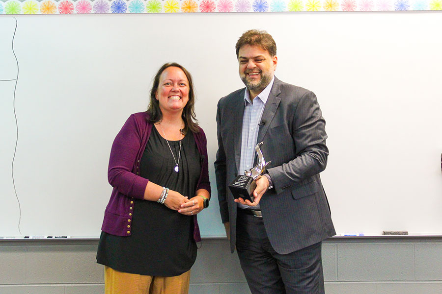 A woman on the left standing next to a man in a suit on the right in front of a whiteboard in a classroom. The man is holding a trophy.