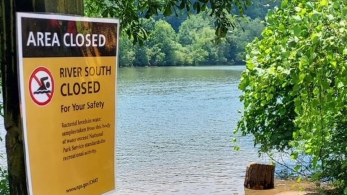 A yellow warning sign beside a river reads “Area Closed. River South closed for your safety.” The sign explains that bacterial levels in the water exceed standards for recreational activity. Trees and calm water are visible in the background.