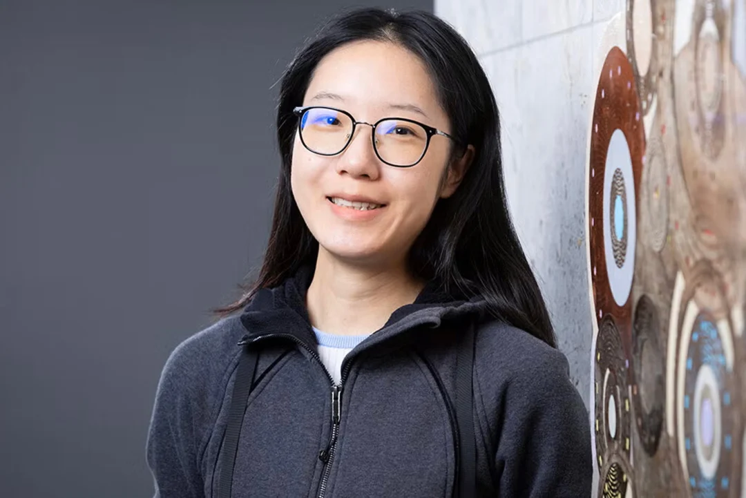 A smiling woman with long black hair and glasses wears a dark gray zip-up hoodie while leaning against a patterned wall.