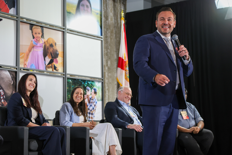 A speaker in a blue suit holds a microphone while addressing an audience onstage, with four panelists seated behind; a Florida state flag and a wall of large photo panels form the backdrop.