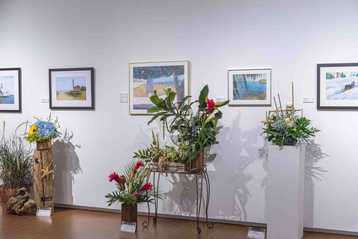 Floral arrangements sit in front of watercolors paintings from the 38th Tri-State Juried Water Media Exhibition at the FSU Museum of Fine Arts.