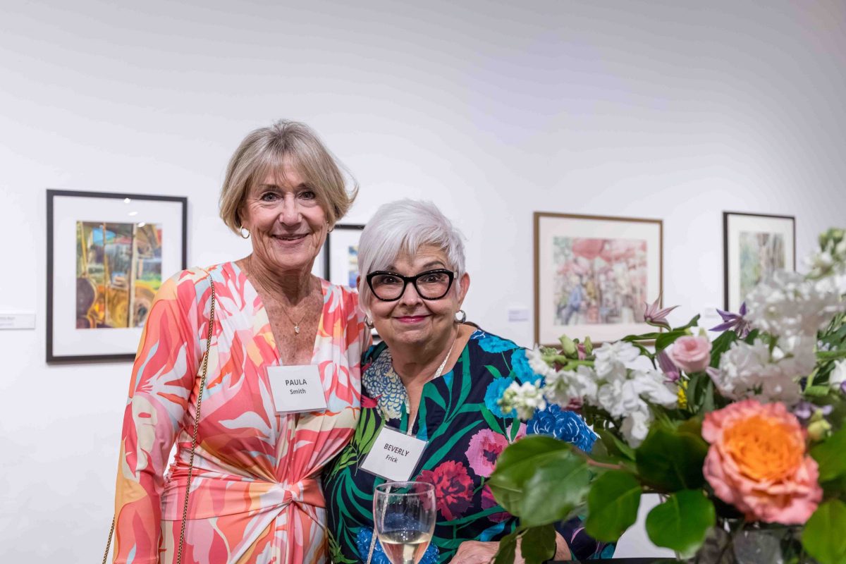 Two women smile among flowers and art.