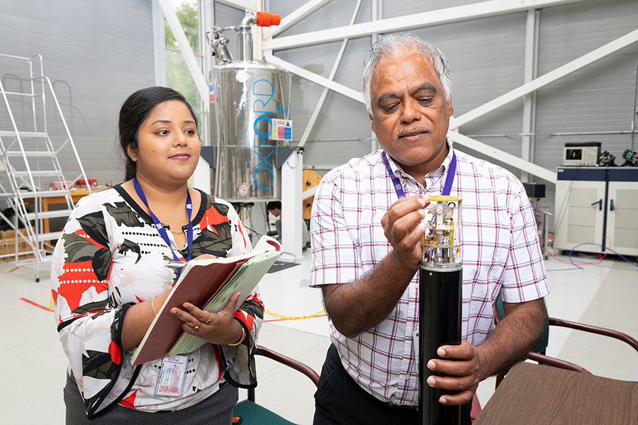 Two researchers stand beside a cylindrical lab instrument, with one holding a notebook and the other examining a small component.