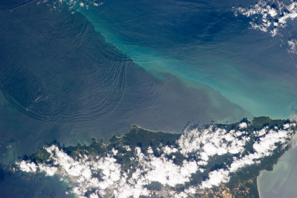 Satellite image of a coastal region showing swirling ocean patterns offshore, with lighter turquoise water near shore and scattered white clouds over a narrow green landmass.