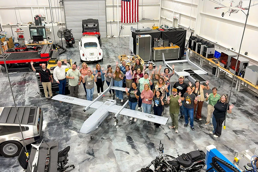 A high-angle, full-color photograph showing a group of people posing and waving inside a spacious engineering hangar, gathered around a grey military drone labeled "USAF."
