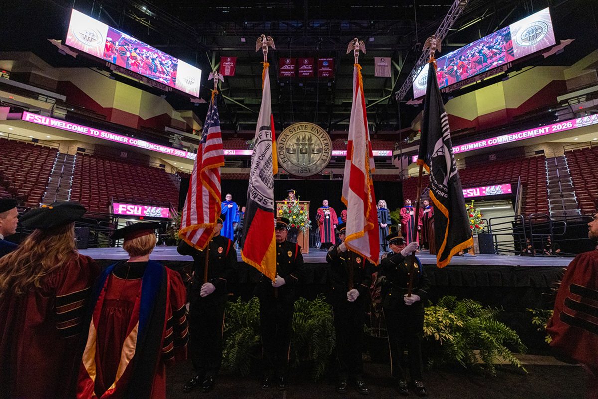 A color guard presents flags on stage in front of spectators and faculty during a commencement ceremony inside an arena.